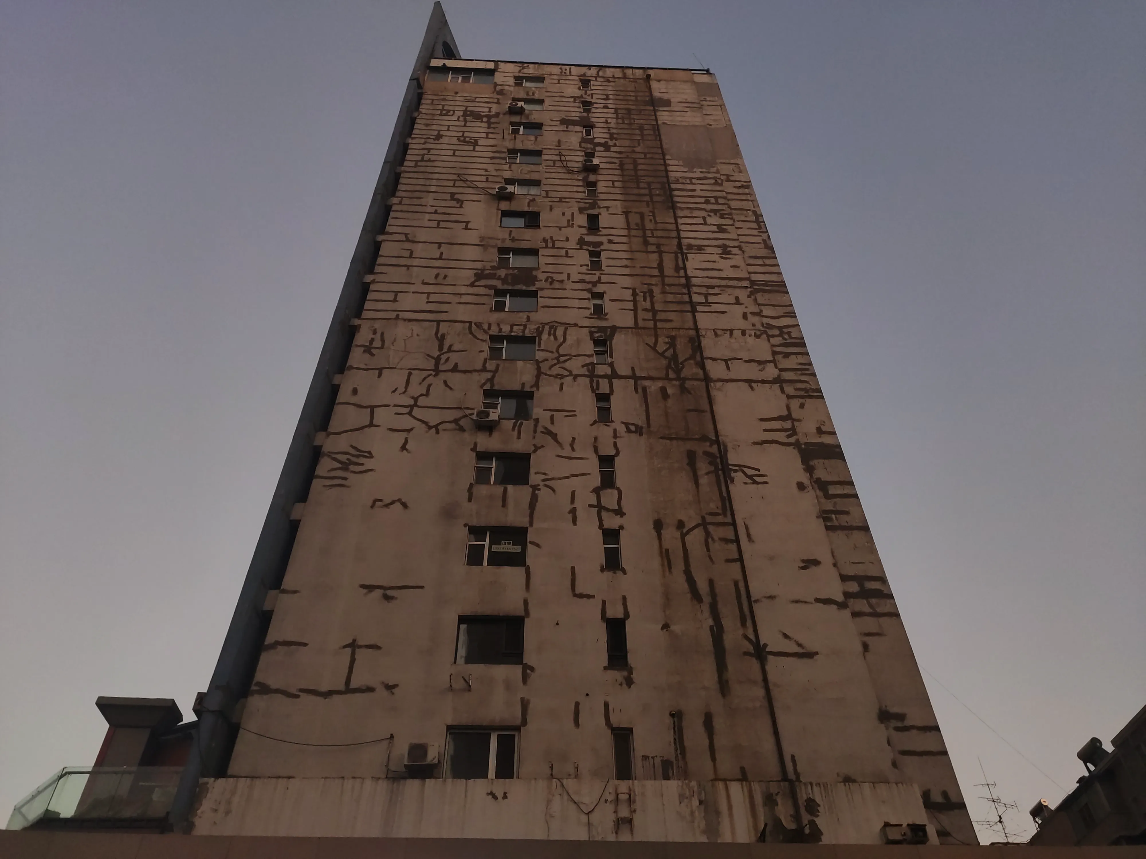 A narrow concrete apartment tower photographed from its base at dusk, its stained façade tapering to a point against a flat violet sky.