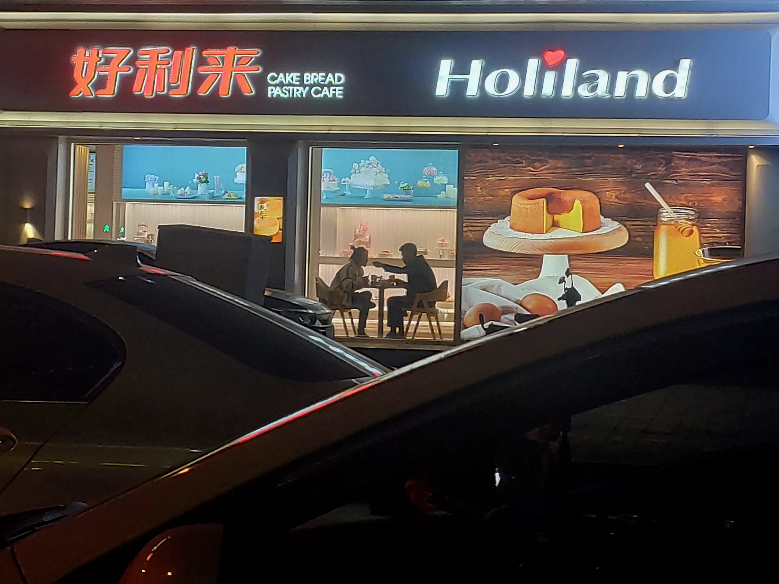 Seen over the roof of a parked car at night: two people sit facing each other at a small table inside a lit bakery, one reaching across.