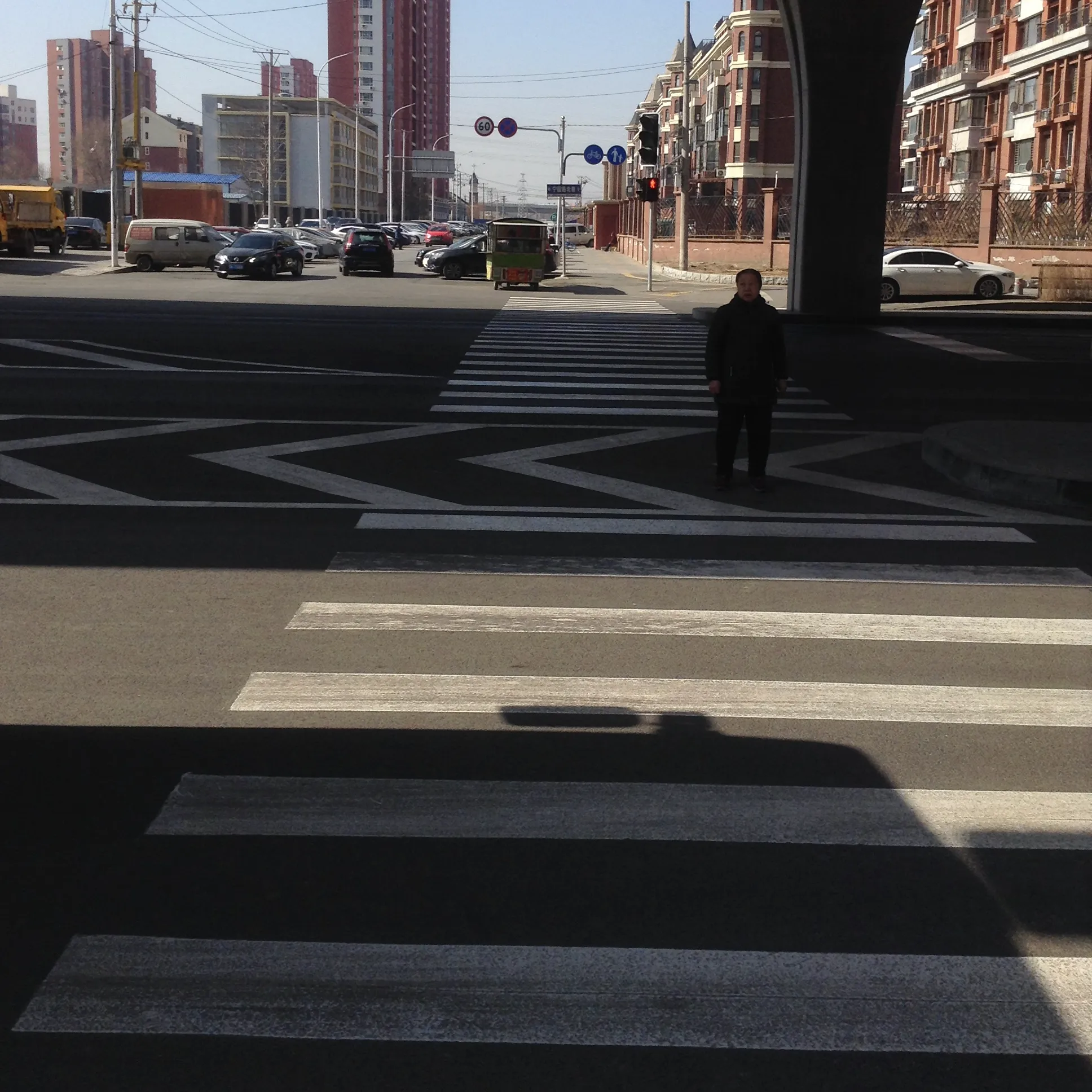 A wide winter crossing painted with chevrons and stripes; a single figure in a dark coat stands alone at the far kerb beneath an overpass column.