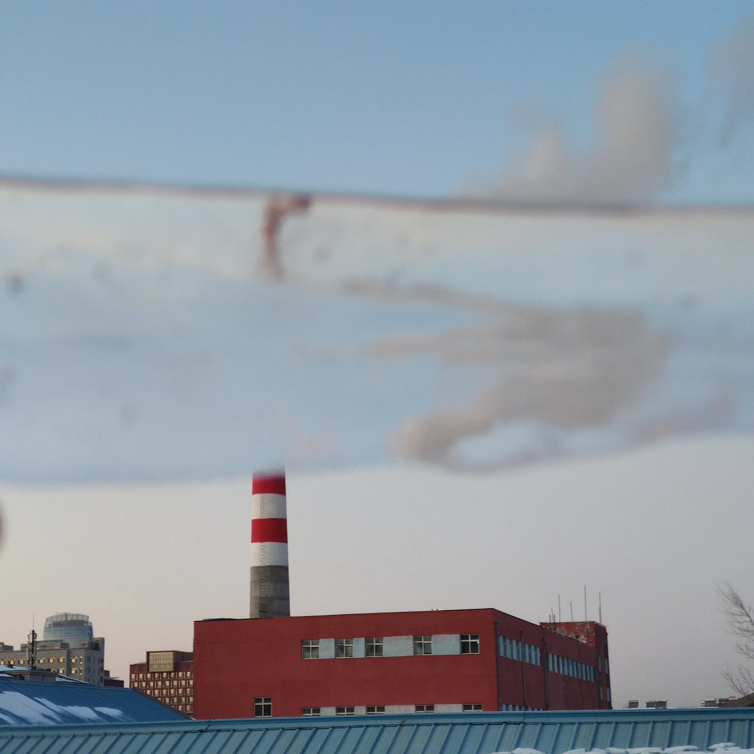 A red-and-white striped chimney behind a red brick plant building, its plume flattening into a long grey band across a pale winter sky.