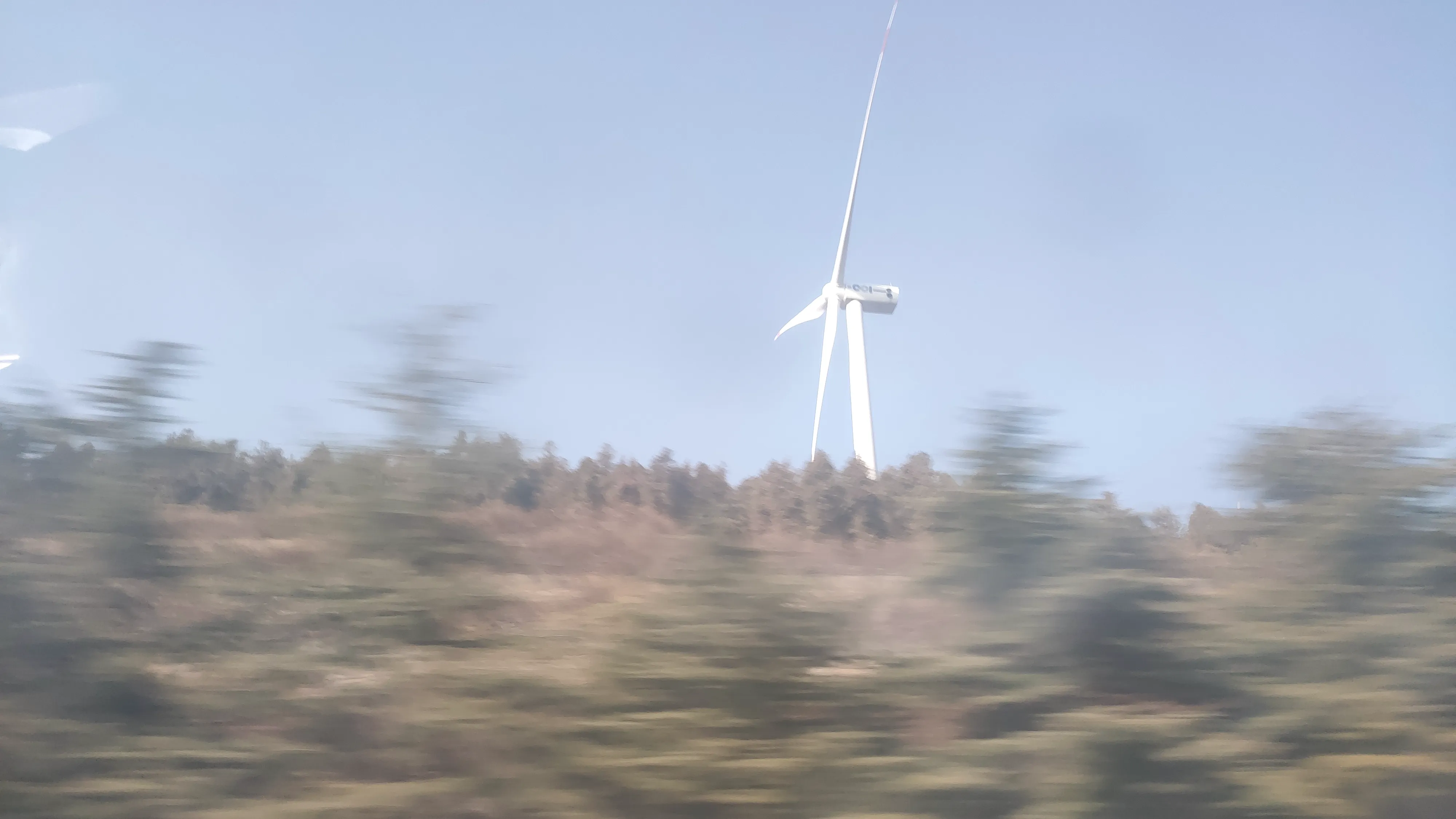 A white wind turbine standing above a blurred hillside of scrub and bare trees, the foreground smeared by motion.