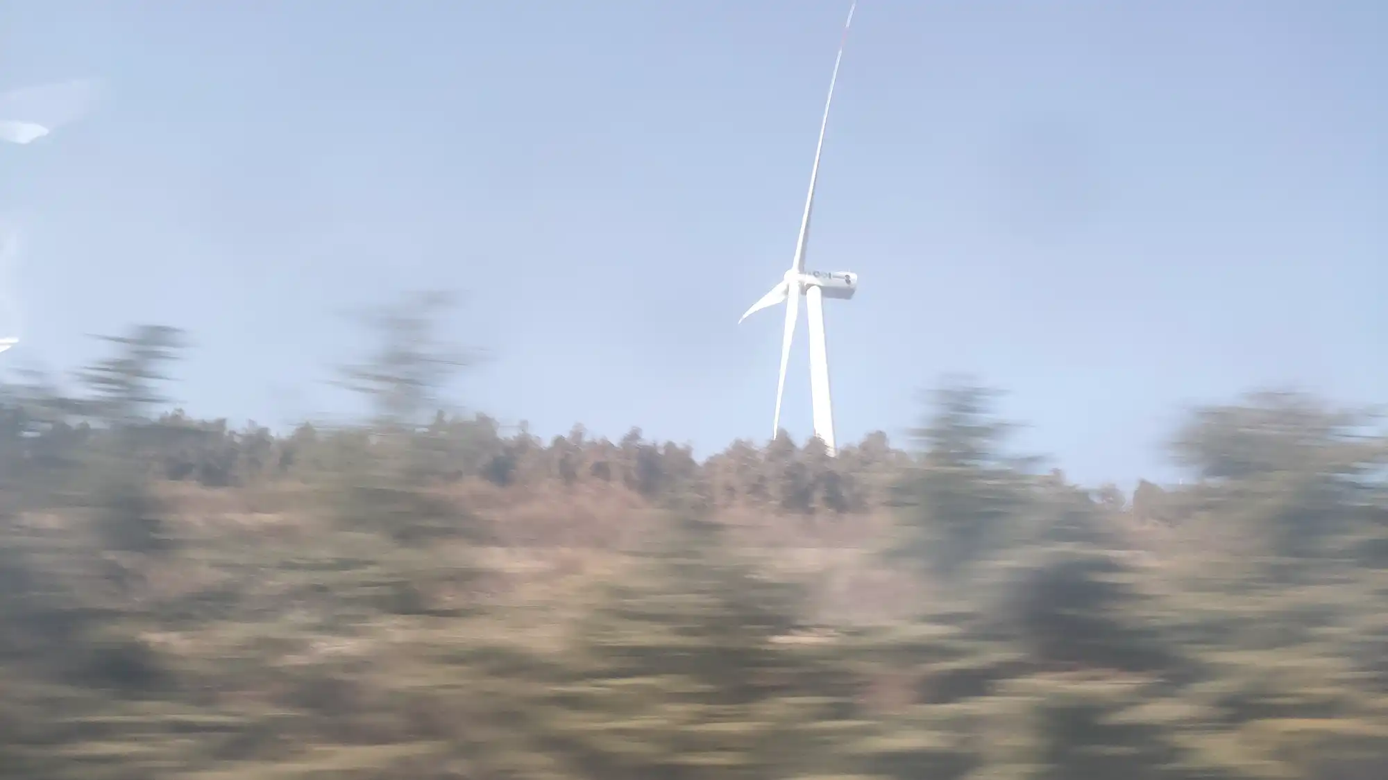 A white wind turbine standing above a blurred hillside of scrub and bare trees, the foreground smeared by motion.