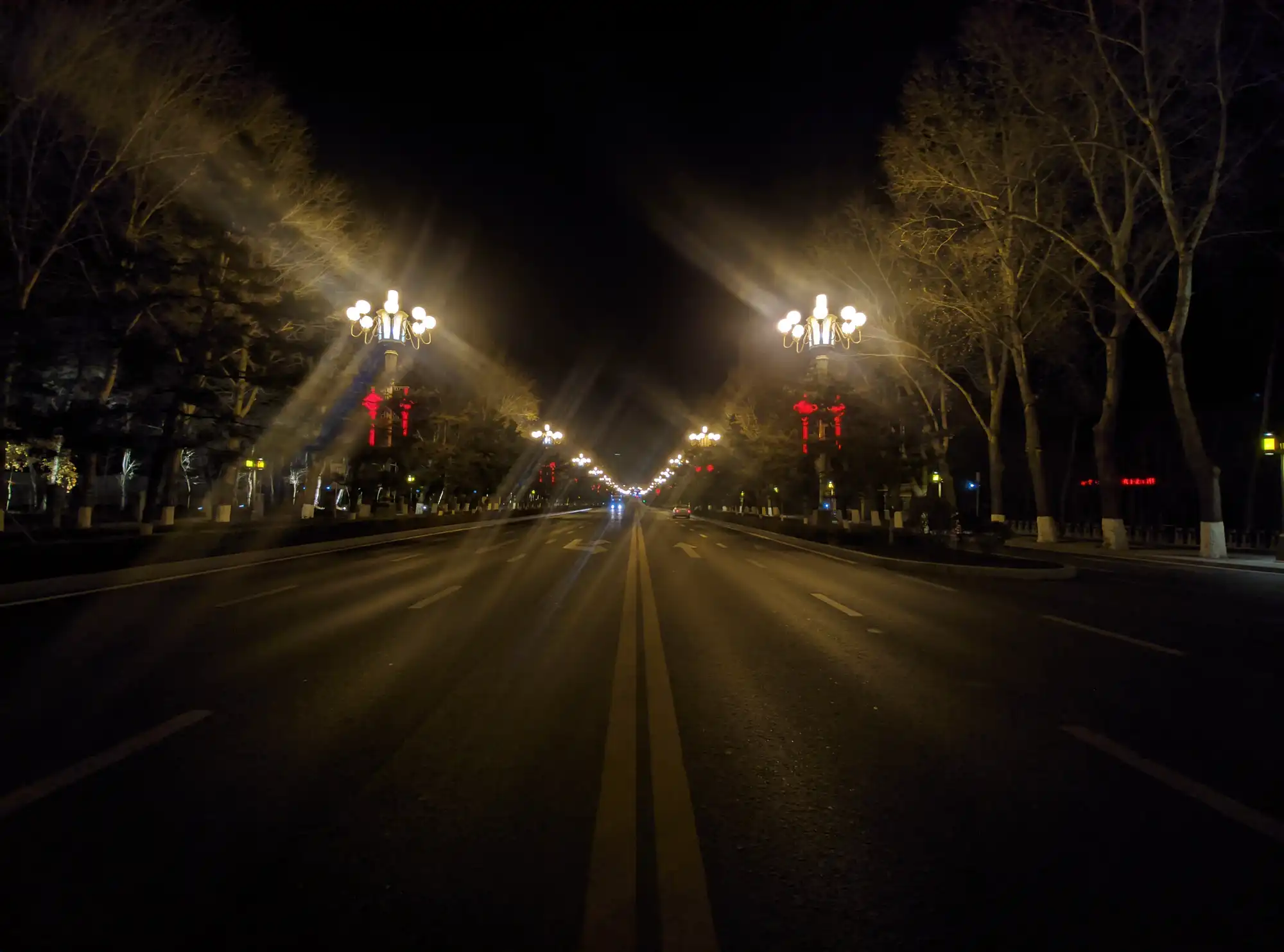 An empty multi-lane avenue at night, bare trees on both sides, ornate street lamps flaring into starbursts and red lanterns hung along the median.