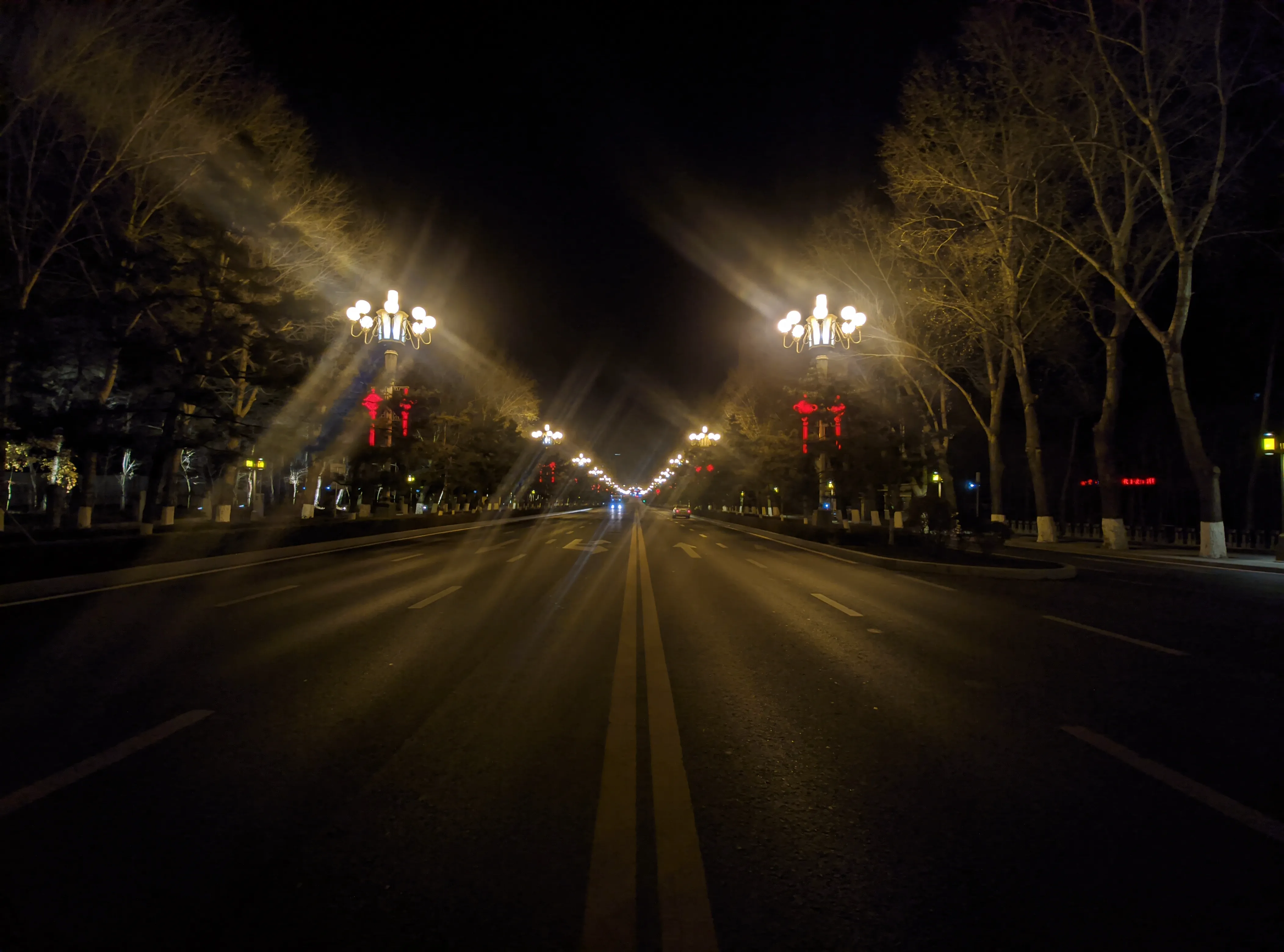 An empty multi-lane avenue at night, bare trees on both sides, ornate street lamps flaring into starbursts and red lanterns hung along the median.