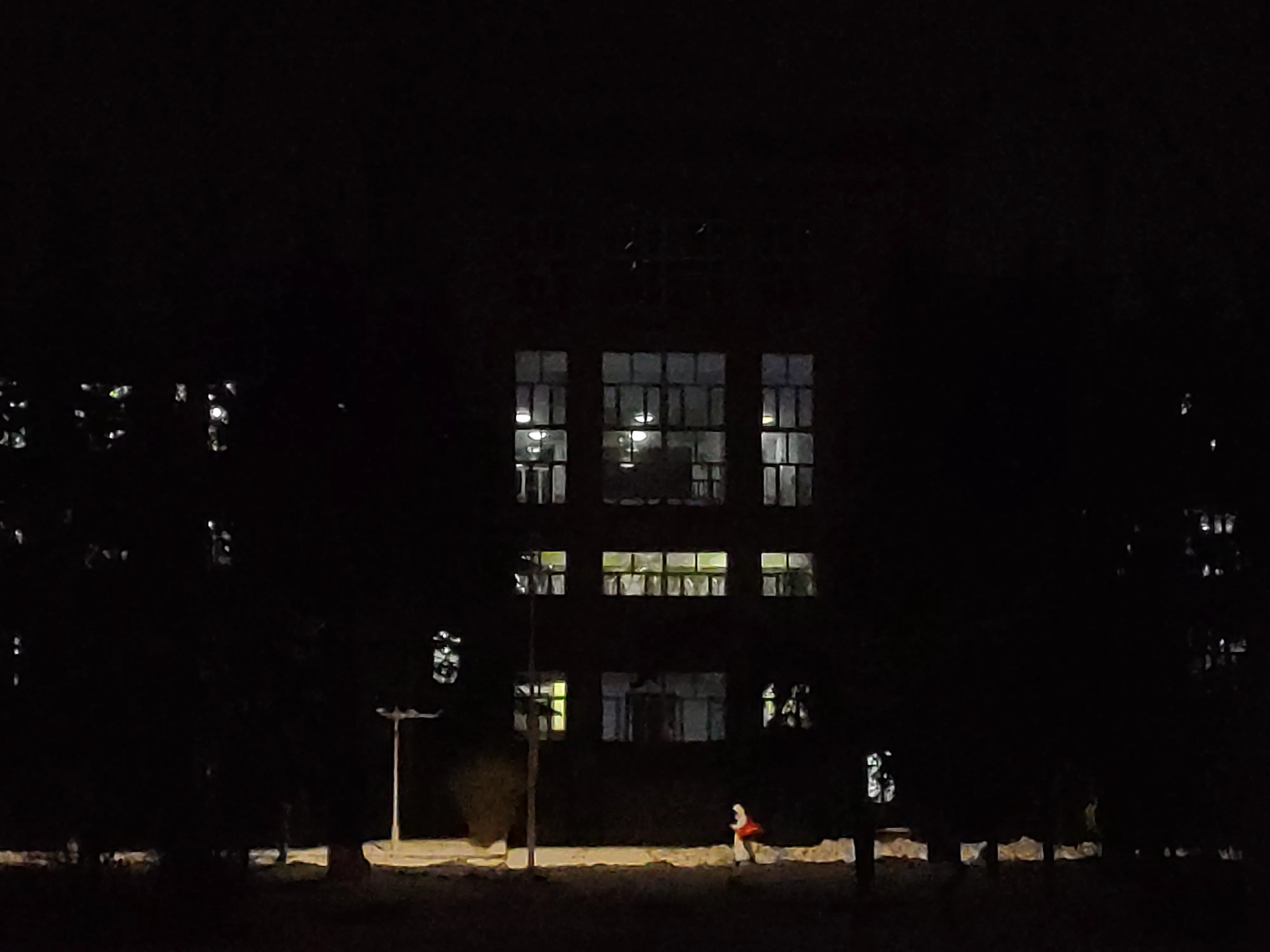 A dark building at night with three bands of lit windows; a small figure in red walks the path below, alone.