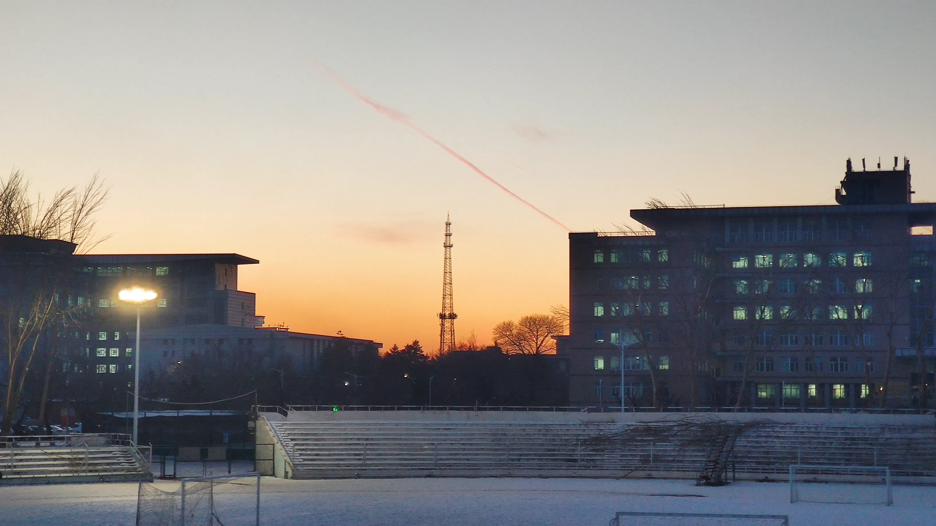 Winter sunset behind a campus: an orange sky crossed by a single contrail, a lattice tower on the skyline, and empty snow-covered stands beside a football pitch.